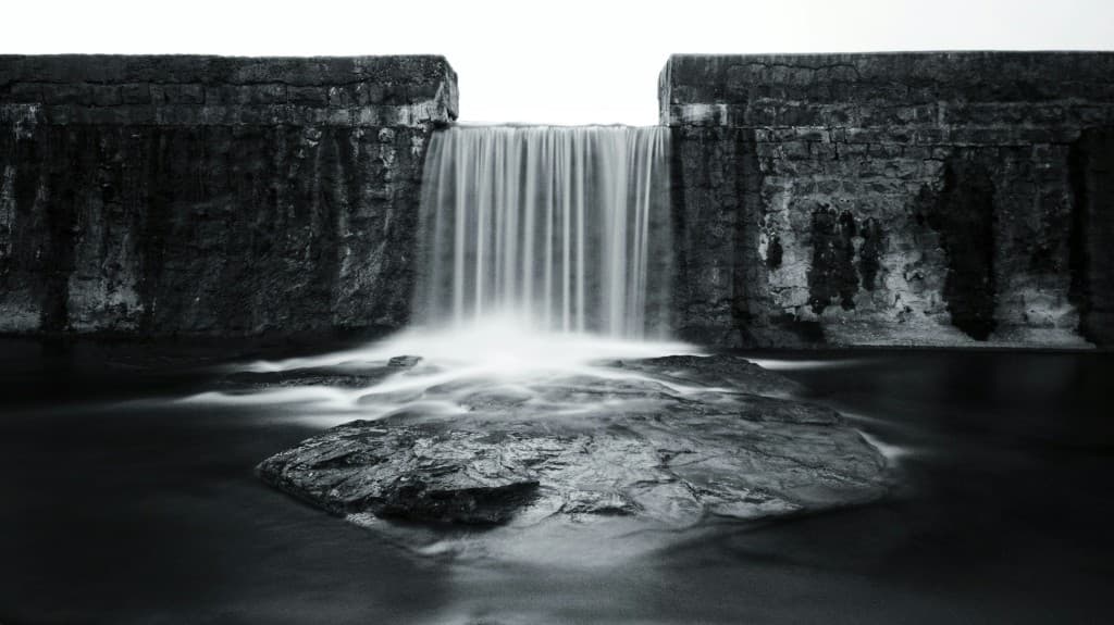 Black-and-white photograph of water spilling through a rectangular notch in a stone wall onto a dark rock